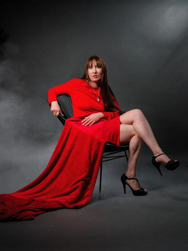 Une femme en robe rouge, assise sur une chaise devant un fond gris